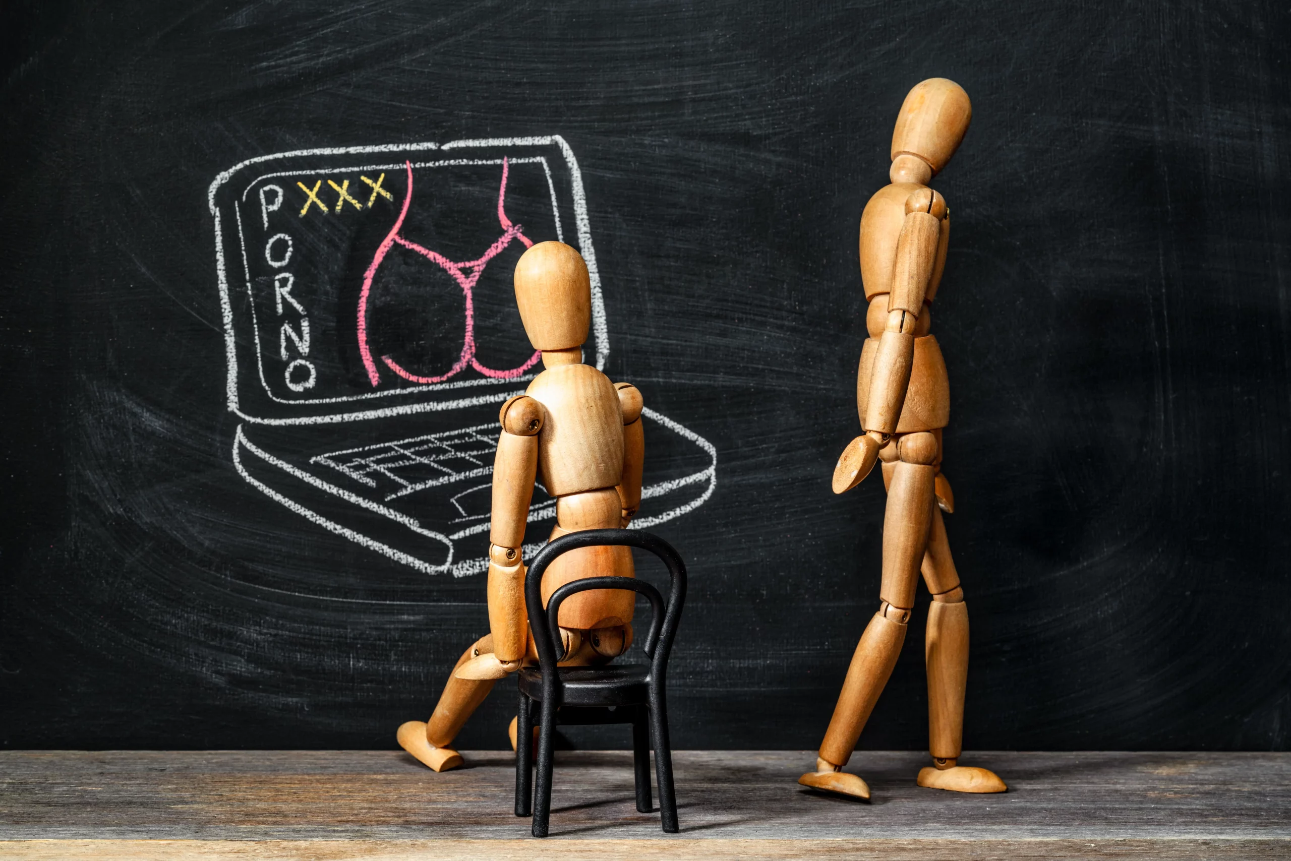 addiction-au-sexe Deux mannequins en bois devant un tableau noir, l'un assis devant un dessin de pornographie sur un ordinateur, l'autre lui tournant le dos et s'éloignant, symbolisant l'isolement et les conséquences des addictions sexuelles.